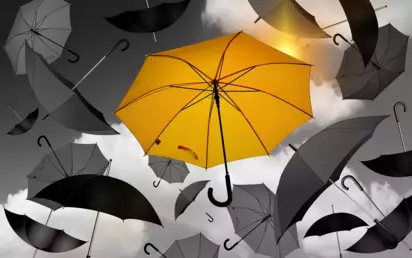 gelber Schirm im Vordergrund, Hintergrund mit vielen grauen Schirmen. Regenschirm als Symbolbild für Schutz.