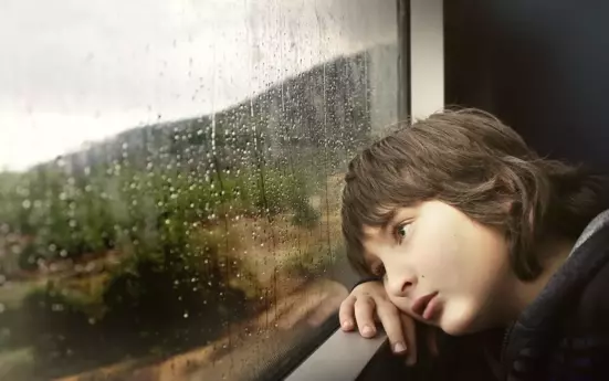 Ein Kind steht vor einem verregneten Fenster und schaut traurig nach draußen.